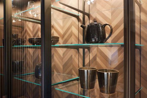 Von grafischer Strenge geprägt ist diese SieMatic Komposition aus Elementen in sterlinggrauem Mattlack, grob texturierter Ceramic und schwarzmatt gebürstetem Aluminium, optisch zusammengefasst von der Stirnwand aus verglasten, beleuchteten Hochschrankvitrinen. Die beleuchteten Griffmulden betonen die horizontalen und vertikalen Linien. © Copyright SieMatic