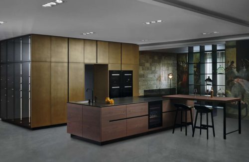 Die Metallfarbe „brass" – eingesetzt bei den Hoch- und Ausgleichsschränken – macht diese Küche einzigartig. Das Furnier „arabica oak" der Kücheninsel und Ansatz- Tischplatte ergänzt den messingfarbenen Metallcolorton perfekt. Edle, kleine Feinheiten wie der Weinkühler zwischen den Unterschränken und Dekorglastüren mit Lichtprofilen runden das Konzept ab.