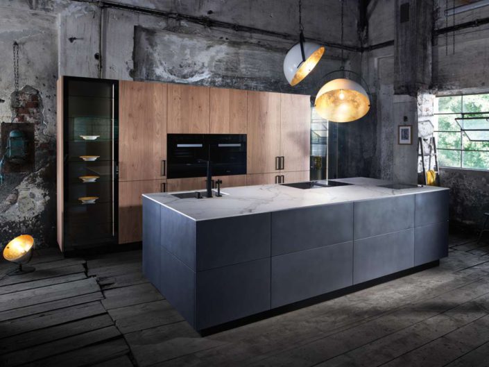 Die Insel in „dark iron" ist das klare Zentrum der Küche. Dazu setzen Hochschränke in Eichenfurnier „white oak" einen stilvollen Kontrast. Der Falttür-Aufsatzschrank bildet das optimale Arbeitszentrum und bietet Stauraum für Küchengeräte. Umfeldmaterialien wie Rahmen, Sockel oder Griffe sind schwarz gehalten. Dekorglas und vertikale Beleuchtung in Hoch- und Oberschränken als bewusst eingesetzte Details vervollständigen das Konzept dieser Küche. Foto: INTUO