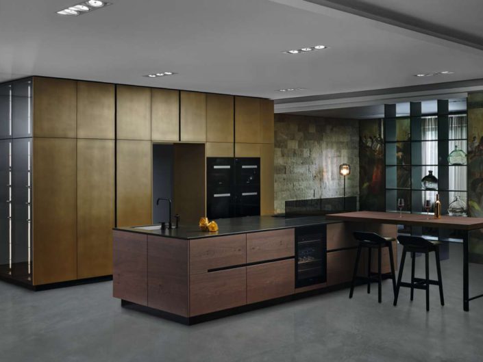 Die Metallfarbe „brass" – eingesetzt bei den Hoch- und Ausgleichsschränken – macht diese Küche einzigartig. Das Furnier „arabica oak" der Kücheninsel und Ansatz- Tischplatte ergänzt den messingfarbenen Metallcolorton perfekt. Edle, kleine Feinheiten wie der Weinkühler zwischen den Unterschränken und Dekorglastüren mit Lichtprofilen runden das Konzept ab.