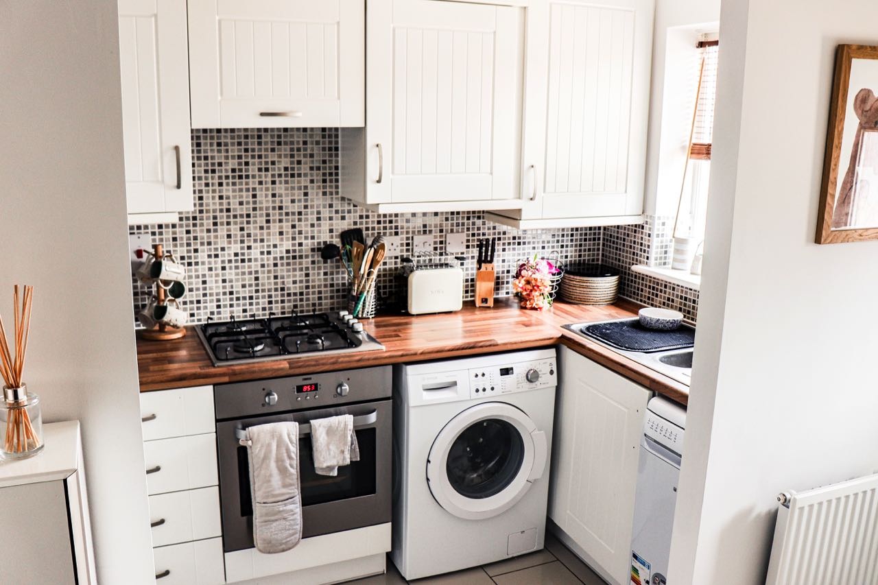 Diese kleine, aber gemütliche Küche kombiniert weiße Küchenmöbel im modernen Landhausstil mit einer warmen Holzarbeitsplatte. Trotz der beengten Verhältnisse findet hier auch noch eine Waschmaschine ihren Platz. Foto: Evy Prentice by unsplash