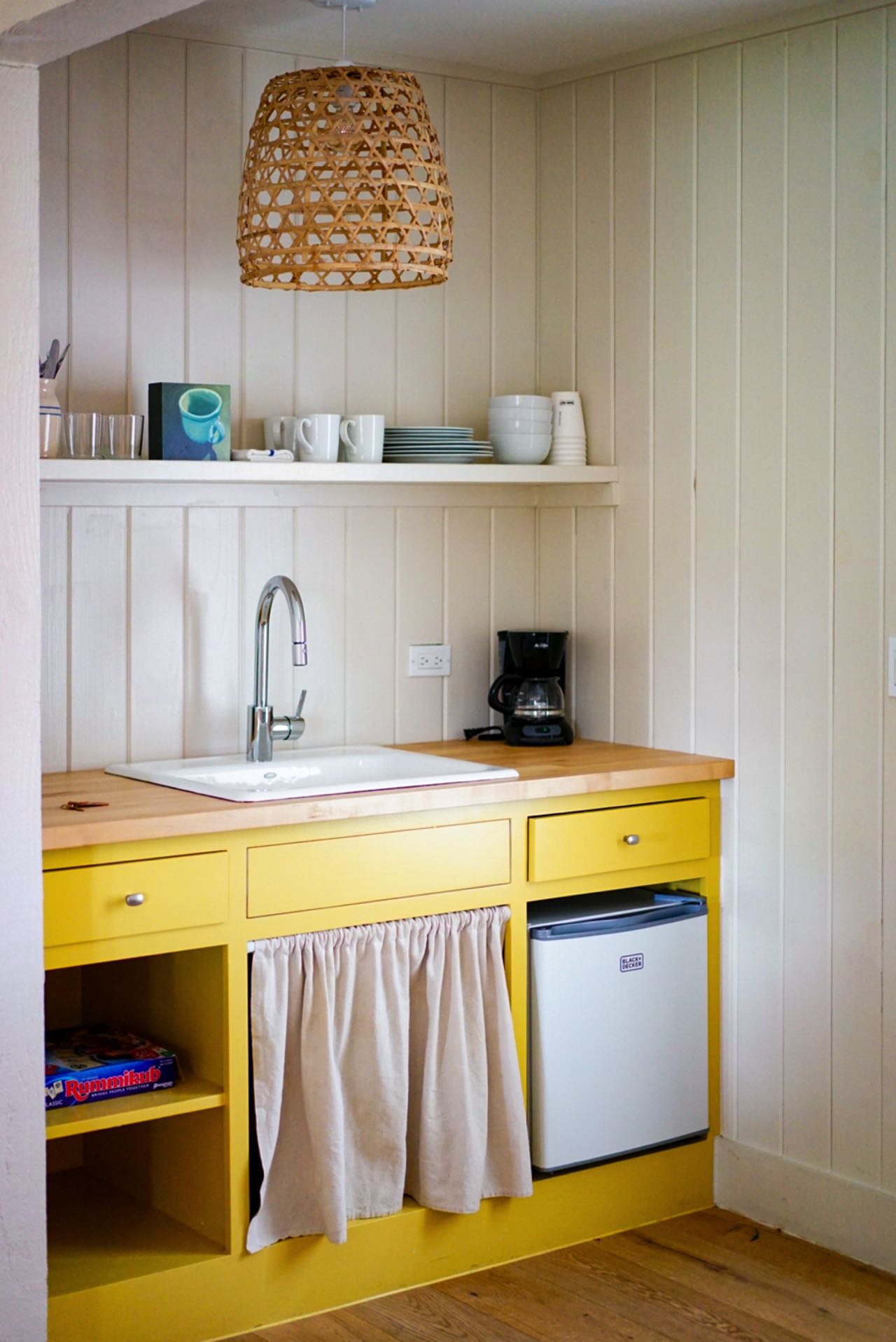 Sonnengelbe Küchenschränke setzen einen lebendigen Kontrast zu den weißen Holzpaneelen an den Wänden. Die natürliche Note der Korbgeflecht-Hängelampe und der Holzarbeitsplatte trifft auf die schlichte Eleganz des weißen Keramikspülbeckens und des einfachen Regals. Foto: Megan Bucknall bye unsplash