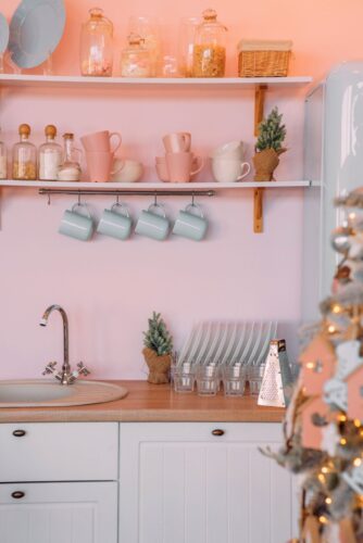 Das zarte Pastellrosa der Wandgestaltung setzt in dieser heimeligen Landhausküche ein sanftes Highlight. Es schafft eine harmonische Verbindung zwischen den weißen Küchenmöbeln und den türkisen Akzenten, wie den Tassen und dem Retro-Kühlschrank. Die liebevoll platzierten Weihnachtsdekorationen unterstreichen die warme Atmosphäre und laden zum gemütlichen Verweilen in der festlichen Jahreszeit ein. Foto: Eugenivy by Unsplash
