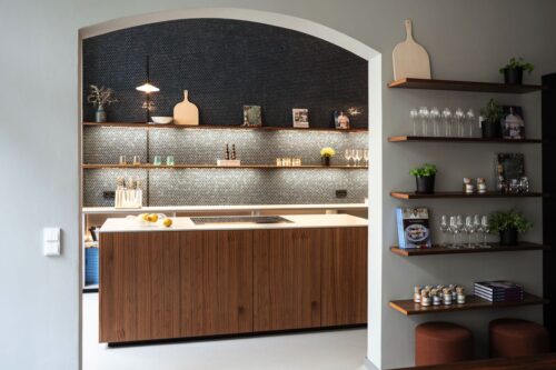 Das beeindruckende Design dieser Küchenwand kombiniert kunstvoll Mosaikfliesen in verschiedenen Farben mit schlichten, warmen Holzdetails. Die Regale, harmonisch in die Wand integriert, präsentieren sowohl Alltagsgegenstände als auch dekorative Akzente und sorgen für eine stimmige Balance aus Ästhetik und Funktionalität. Alles von next125.