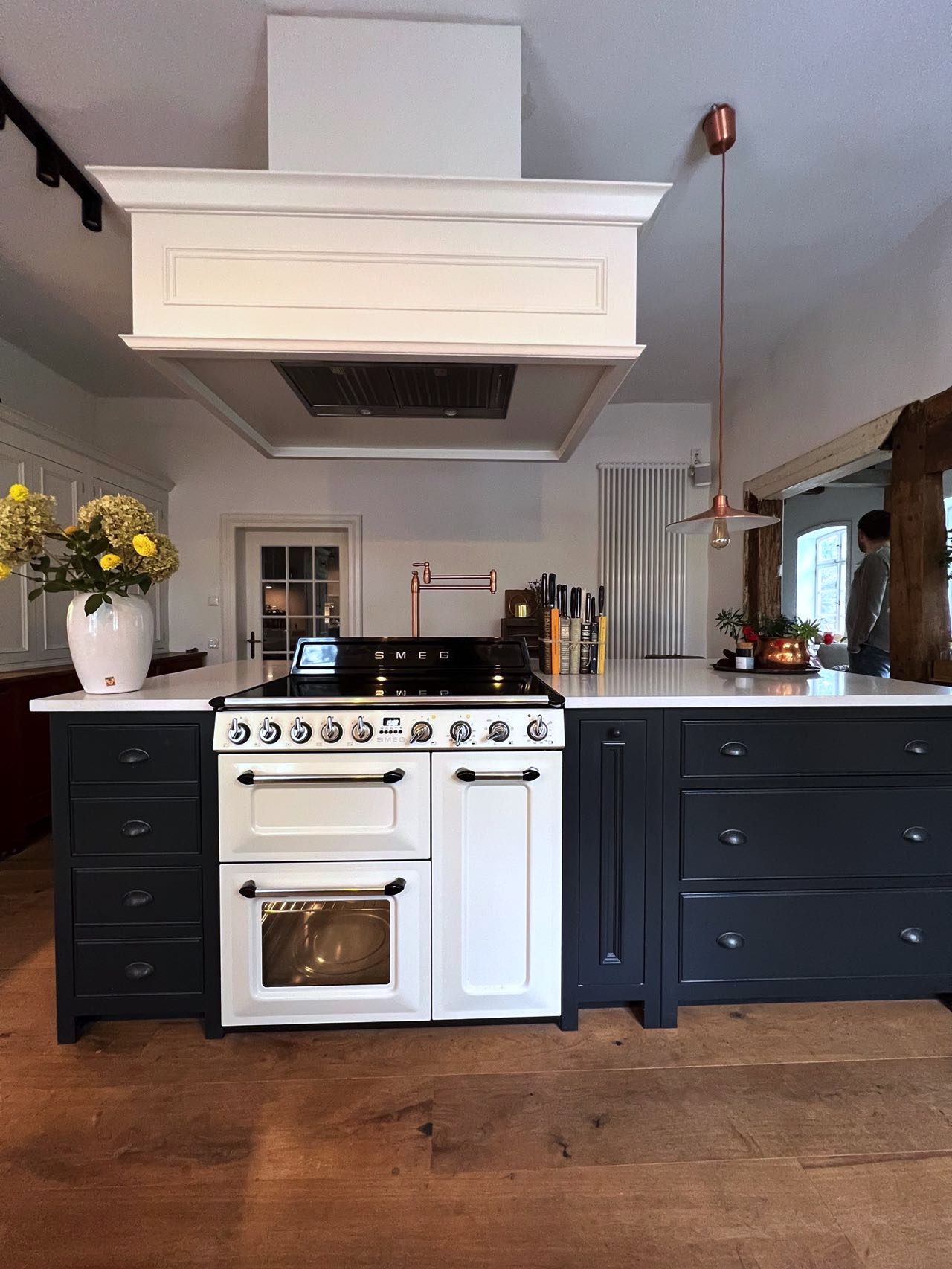 Im Zentrum der Küche beeindruckt die Kücheninsel mit einem stilvollen, weißen SMEG-Rangecooker und einer ungewöhnlichen Dunstabzugshaube, die für ein offenes Raumgefühl sorgt.
