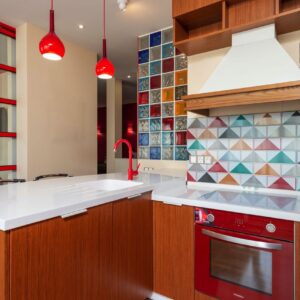 Mutig wurden hier rötliche Küchenfronten aus Holz mit roten Details kombiniert. Diese Farbe findet sich auch in den Glasbausteinen sowie der kunterbunten Fliesenrückwand. Ruhe bringt nur die weiße schlichte Arbeitsplatte mit passender Spüle in die Szenerie. Foto: Max Rahubovskiy | Pexels