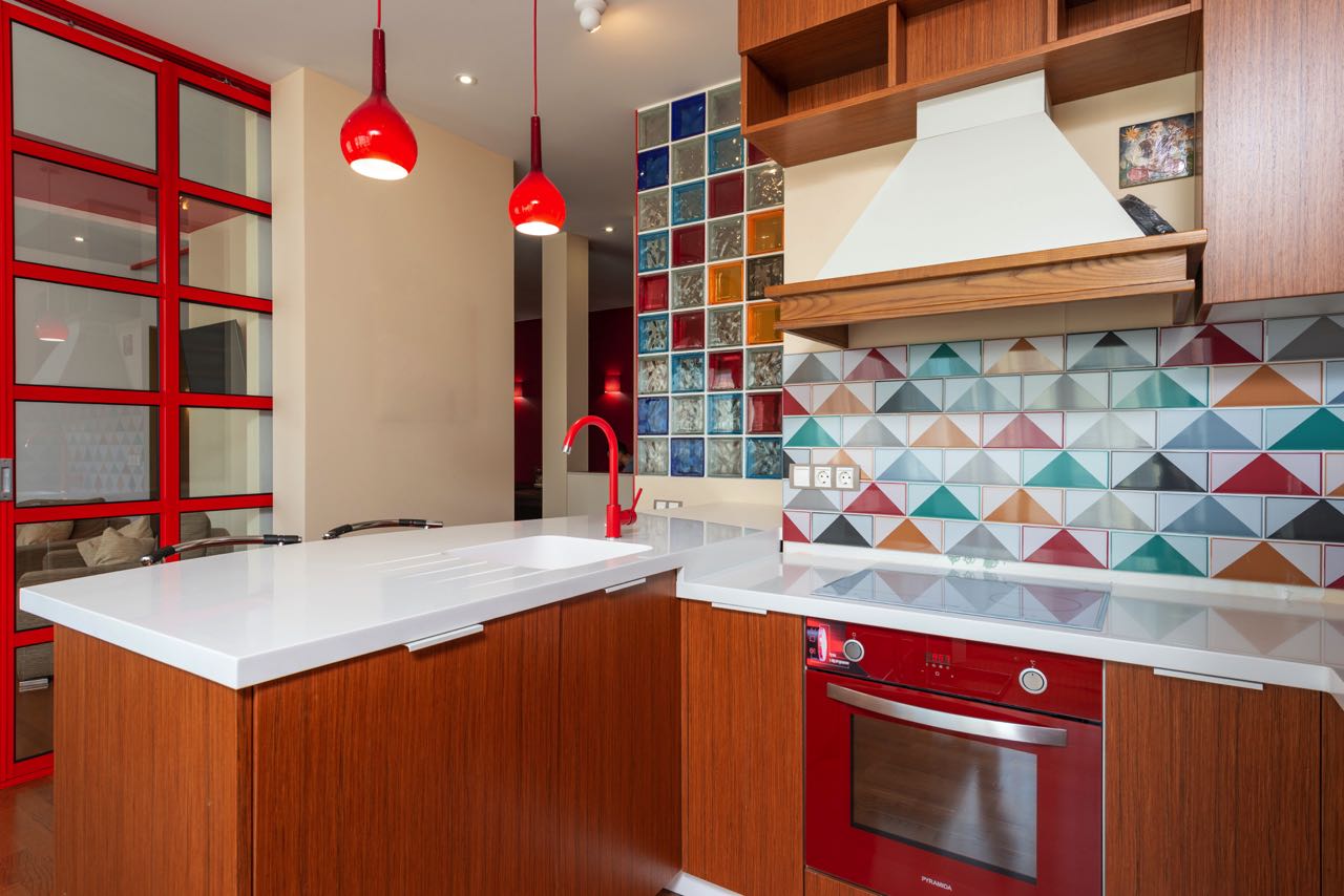 Mutig wurden hier rötliche Küchenfronten aus Holz mit roten Details kombiniert. Diese Farbe findet sich auch in den Glasbausteinen sowie der kunterbunten Fliesenrückwand. Ruhe bringt nur die weiße schlichte Arbeitsplatte mit passender Spüle in die Szenerie. Foto: Max Rahubovskiy | Pexels