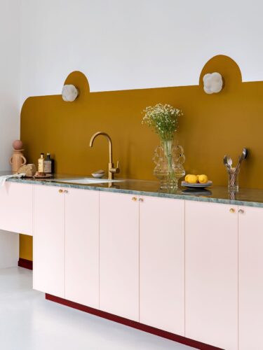 Wer sich gegen Regale oder Schränke an der Wand entscheidet, hat neu gewonnenen Platz für eine hübsche Gestaltung. In diesem Fall ist das eine kunstvoll bemalte, farbige Wand mit Lampen. Küche von Plum Living.