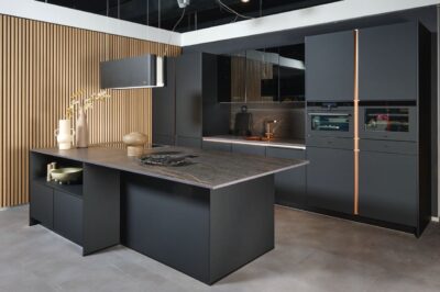 Auf den ersten Blick wirkt die Küche eher streng und opulent mit dunklem Marmor und dunklen Fronten. Das Kupfer verstärkt den edlen Look. Aufgebrochen wird die Strenge durch die helle Holzwand, die eine Spur Unkompliziertheit in den Raum bringt. Von Impuls Küchen.