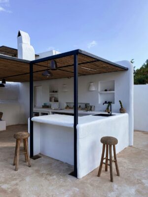 Diese Outdoor-Küche vereint Funktion und Stil in einem schlichten, aber ansprechenden Design. Die weiß verputzte Struktur strahlt in der Sonne und erinnert an die klassische Architektur des Mittelmeerraums. Die in die Wand integrierten Regale bieten praktischen Stauraum, während die schwarzen Metallelemente einen modernen, industriellen Akzent setzen. Die robusten Holzstühle mit gewebten Sitzen fügen eine rustikale Note hinzu, die das Gesamtbild erdet. Unter dem Dach aus natürlichen Materialien ist die Küche teilweise vor den Elementen geschützt, während die eingelassenen Leuchten in der Rückwand für eine stimmungsvolle Beleuchtung der Arbeitsfläche bei Dämmerung oder Dunkelheit sorgen. Foto: Jubeo Hernandez by unsplash