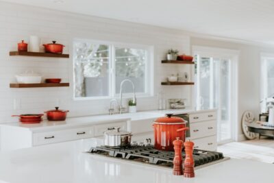 Helle Küche mit Akzenten in Rot – Eine stilvolle, moderne Küche mit weißen Arbeitsflächen und Bodenfliesen sowie roten Kochutensilien als Hingucker. Die großen Fenster lassen viel Tageslicht herein. Foto: Unsplash, Becca Tapert.