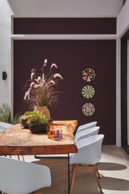 Der Farbton Mocha Mousse ist die Pantone Farbe des Jahres 2025. Sie verschönert die Wand und strahlt Wärme aus. Die Wandfarbe ist von Little Greene. Die weißen Stühle bilden dazu farblich einen gekonnten Kontrast. In Sachen Stil schmeicheln die Design-Stühle auch dem eher robusten Tisch aus einer Baumscheibe. Die Accessoires verbreiten Sommer-Feeling.