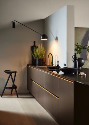 Der puristische Look kann auch mit dunklen Farben in einer kleinen Küche umgesetzt werden. Die offene Raumlösung ist dafür ideal. Durch die Akzentlampe wird sie gezielt ins rechte Licht gesetzt werden. Küche „Soft Lack“ in der Farbe „Sepiabraun“ von Nolte Küchen.