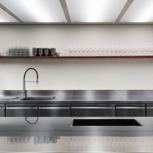Grober Stein kontrastiert zur cleanen Arbeitsfläche aus Edelstahl. Die zweite Zeile ist komplett aus Edelstahl, sowohl die Fronten als auch die Arbeitsplatte. Das harmoniert gut und wirkt optisch aufgeräumt. Von Quadro Design. 