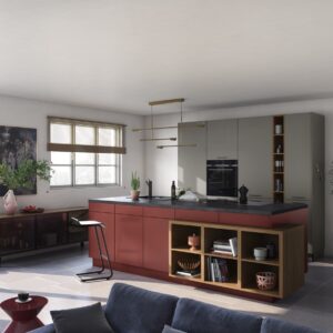 Die Kücheninsel fällt sofort ins Auge. Sie begeistert mit dem attraktiven Rotton. Er bildet einen gelungenen Kontrast zum Grau. Holz- und Golddetails schmeicheln dem Ganzen. Die Möbel sind von Küche & Co.