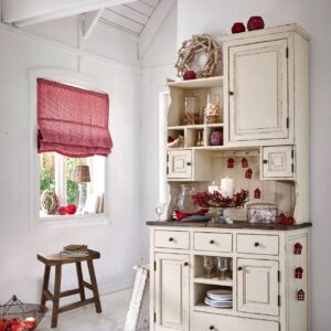 Tradition mit modernem Twist. Der beigefarbene Küchenschrank im Shabby-Look verbindet sich mit zurückhaltendem Rot zu einem warmen, festlichen Landhaus-moment. Modern ist das Arrangement auf der Etagere und die Lichterkette aus klei-nen Häusern. Alles von Loberon.