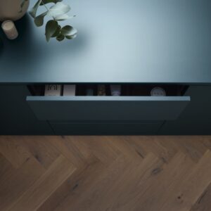 Das Material „Rauvisio Crystal“ von Rehau ist ein smartes Glas, das im Bereich Arbeitsflächen genutzt wird. Das smarte Glas ist bruchfester und leichter als Echtglas. Natur und Technologie verschmelzen hierbei zu einem ästhetischen Gesamtkonzept. Die Farbwelt dieser Küche ist geprägt von klaren, modernen Strukturen. Ein dunkles Marineblau mit petrolfarbenen Untertönen ist der Farbton „Onda“.