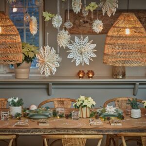 Über dem Tisch schweben filigrane Papiersterne in Weiß, die für winterliche Leichtigkeit sorgen. Die selbst gestalteten Platzsets sind eine kreative Idee. Frische Weihnachtssterne in zarten Cremetönen sowie kleine Zweige und Zapfen ergänzen den handgemachten Look und machen die Dekoration warm, kreativ und wunderbar persönlich.