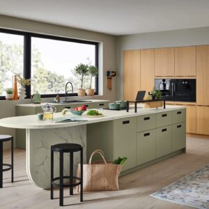 Eine sehr wohnliche Planung in Eichenfurnier und der Trendfarbe Jadegrün zeigt sich bei der großzügigen Küche. Die verlängerte Arbeitsplatte der großen Insel mit ihrer extravaganten, abgerundeten Abschlusskante lädt zum Verweilen und Genießen ein und setzt ein echtes Statement.