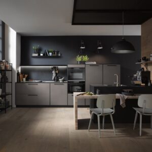 Der Industrial-Stil wird durch Holz ergänzt. Die Fronten in „Perlgrau“ sind schlicht, dennoch erscheinen sie nicht zu kühl. Wohnlich wirken auch die Griffe. Verstärkt wird alles von den Lampen und der Gestaltung des restlichen Raumes. Küche von Häcker.