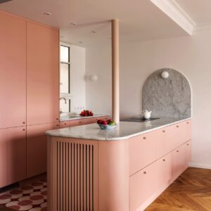 Die eigentlich gerade Küche wurde mit runden Akzenten versehen und wird dadurch zu etwas ganz Besonderem. Dazu zählt die Wandfläche aus Marmor, die Säule und vor allem der angesetzte Schrankbereich mit Lamellen. Im Ton „Blush“ von Plum Living.