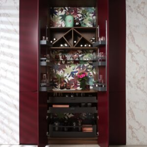 Im Hintergrund kommen Blumen und Blätter optimal zur Geltung und setzen das Möbelstück in Szene. Der Barschrank steckt hinter den farbigen Schranktüren. Er besteht aus mehreren modularen Regalsystemen, die nach eigenen Vorstellungen bestückt werden können, von Schmidt Küchen.