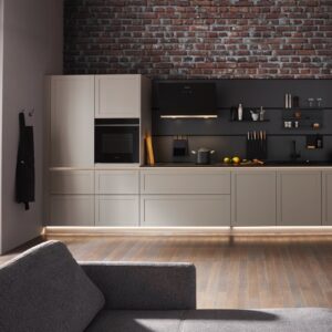 Greige kann auch rustikal. Das beweist diese Küche mit grober Steinwand, industriellen Fenstern und schwarzer Multifunktionsrückwand. Die Küche mit klaren Kassetten-Fronten von Global Küchen mildert den Industrial-Style gekonnt ab.