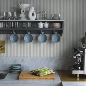 Die feine Streifentapete „Mille Stripe“ in Blau von Boråstapeter bringt eine softe Anmutung in die Küche. In Verbindung mit hellgrauen Fronten, offenem Regal und Keramikgeschirr entsteht ein wohnlicher Look, der Landhausdetails modern aufleben lässt.