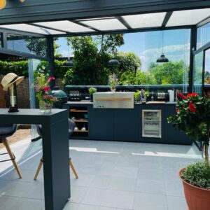 Die Outdoor-Küche beweist, wie sich Kochen im Freien ganzjährig denken lässt. Unter Glas geschützt, entsteht hier ein wetterunabhängiger Kochplatz. Er kann zu allen Seiten hin geöffnet und geschlossen werden kann. Integriert sind Geräte, Stauraum und ein Barbereich. So kann die Küche rund ums Jahr genutzt werden. Küche von Unikaad mit Keramikgrill von Monolith und Gasgrill von Flammkraft.