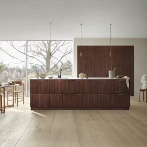 Die Küchenserie „Bordo Nordic Glow“ von Kvik setzt auf dunkles Holz mit markanter Maserung und kombiniert es mit einem hellen Holzboden und Messinggriffen. Die großzügige Insel bildet den Mittelpunkt des Raumes und unterstreicht den klaren skandinavischen Stil. So entsteht eine Atmosphäre, die gleichermaßen warm und zeitlos erscheint.