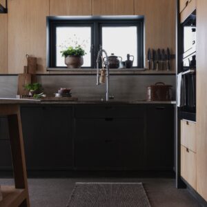 Der dunkle Teppich von Pappelina fügt sich gut in das Bild des tollen Terrazzo-Bodens ein. Beide kontrastieren gekonnt zu den hellen Holzfronten. Schwarze Akzente an Fenstern, Leuchten und Armaturen geben der Küche zusätzliche Kontur. Dadurch wirkt der Raum zeitgemäß und behaglich.